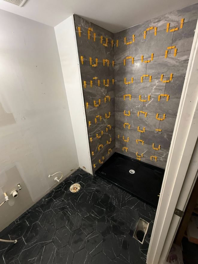Shower walls being tiled in large grey porcelain with leveling clips over a black hexagon floor